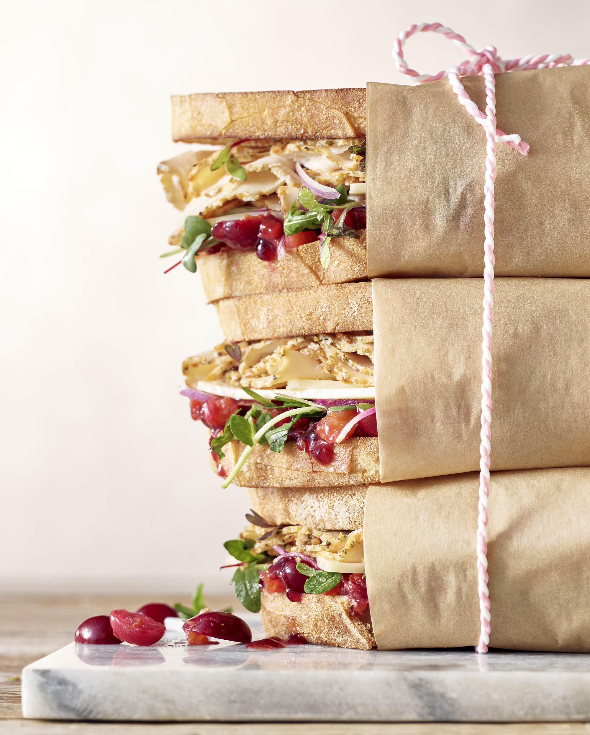 Stack of three turkey sandwiches on white bread wrapped in brown butcher paper with a pink string tying them together.